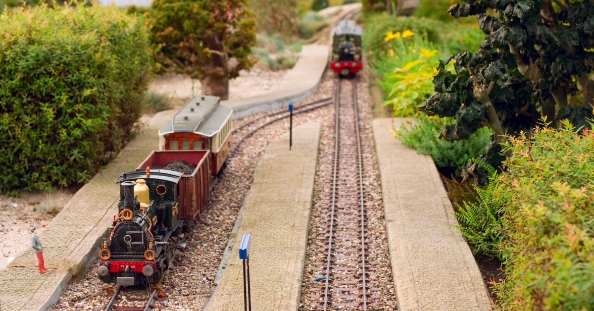 A small model railway with two model trains traveling down different parts of a splitting railroad track surrounded by greenery.